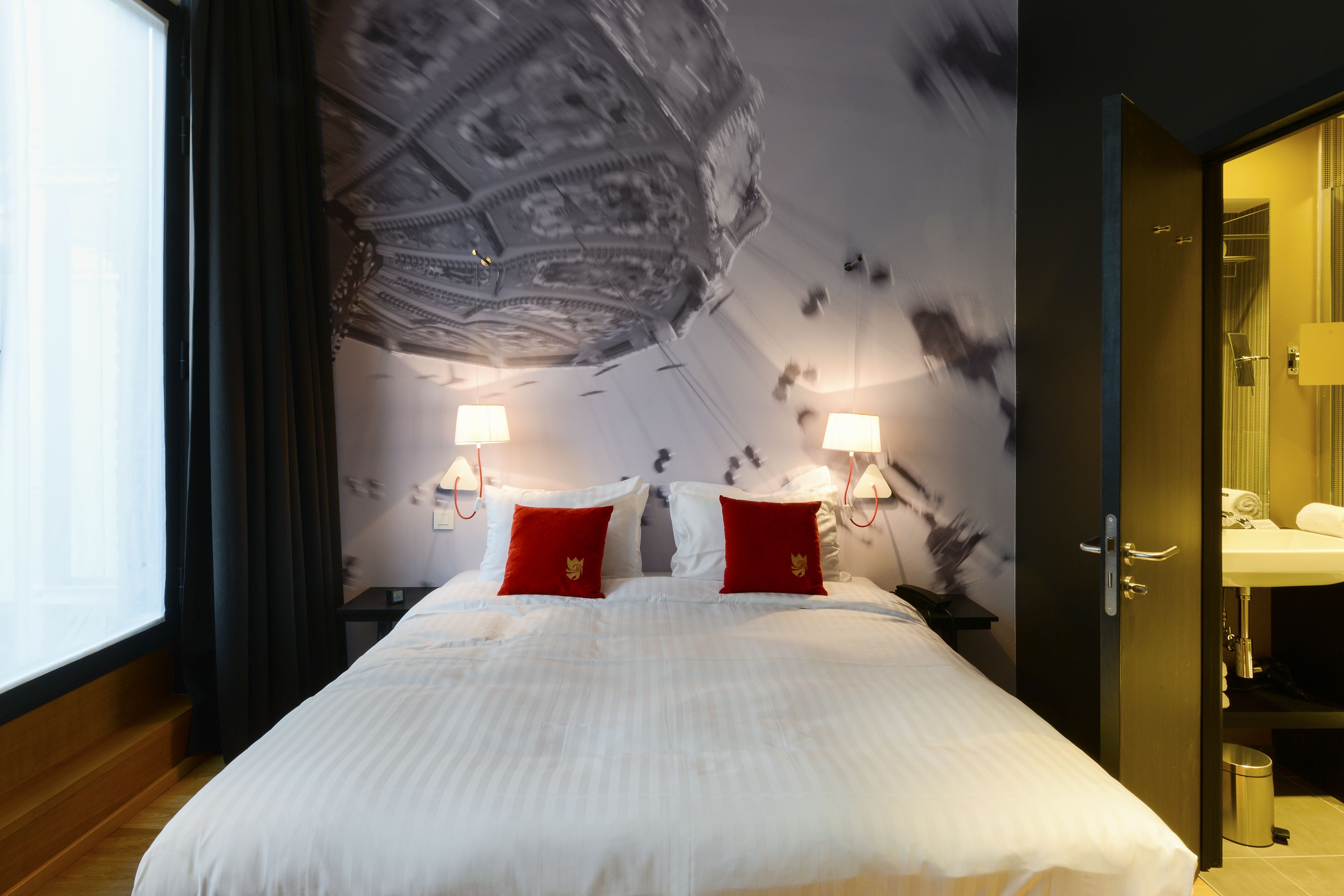 Quarto de hotel com uma cama grande, duas almofadas vermelhas, uma mesa de cabeceira, um abajur fixado na parede e um banheiro visível por uma porta aberta.