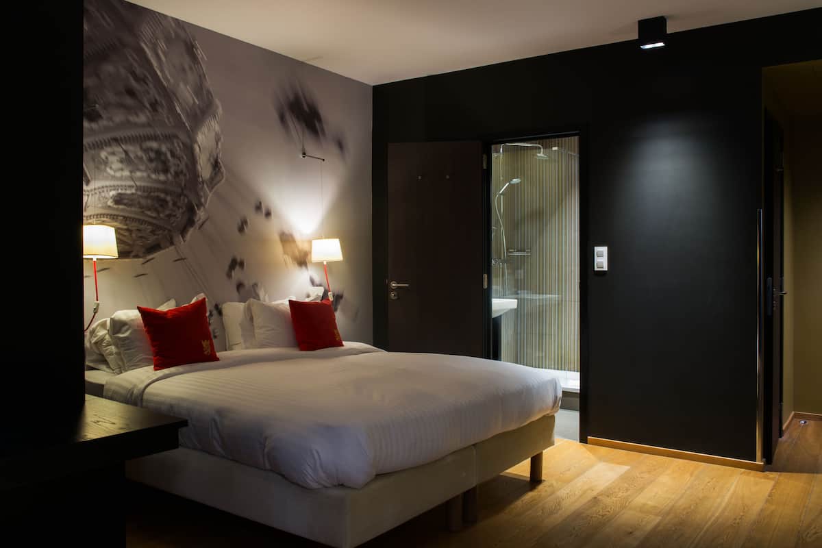 A modern hotel room with a large bed, two bedside tables with lamps, a large abstract wall mural, and a glass shower area.