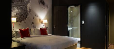 Une chambre d’hôtel moderne dotée d’un grand lit, de deux tables de chevet avec lampes, d’une grande fresque murale abstraite et d’une douche en verre.