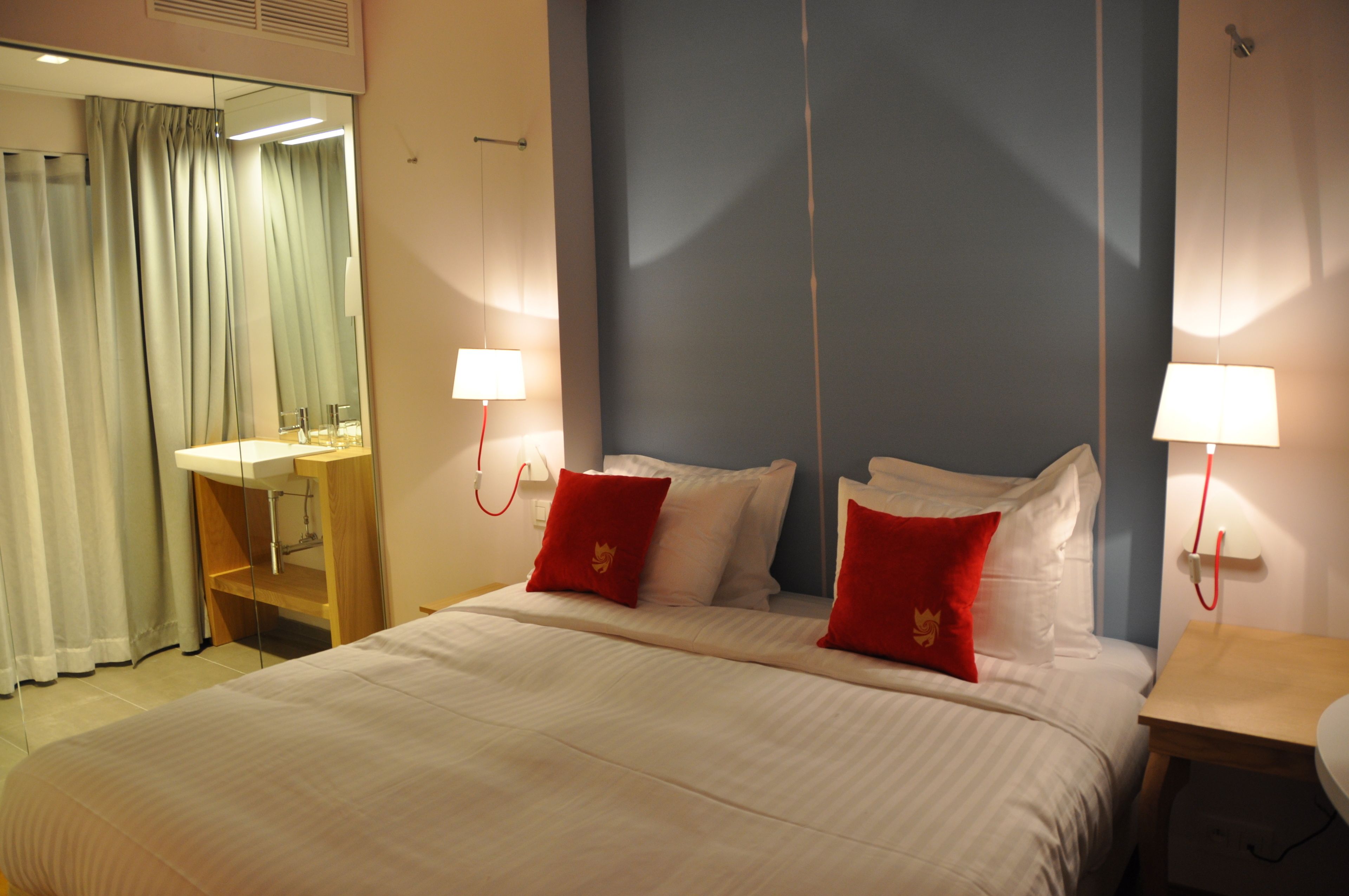 Une chambre d’hôtel avec un grand lit, deux oreillers rouges, une table de chevet avec une lampe, un miroir et une salle de bain avec un lavabo et des rideaux.