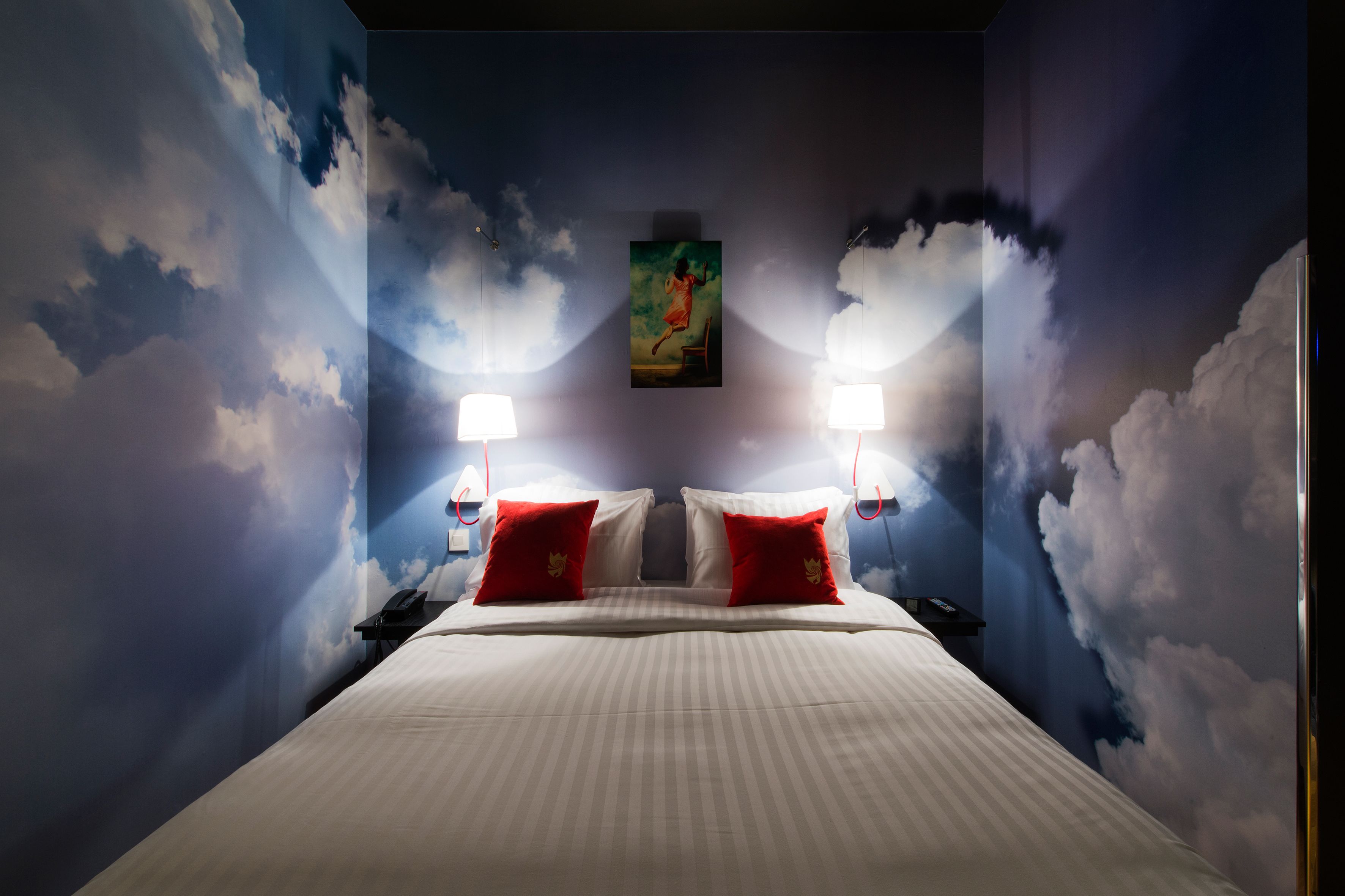 Een hotelkamer met een bed, twee rode kussens, een schilderij aan de muur en twee wandlampen.