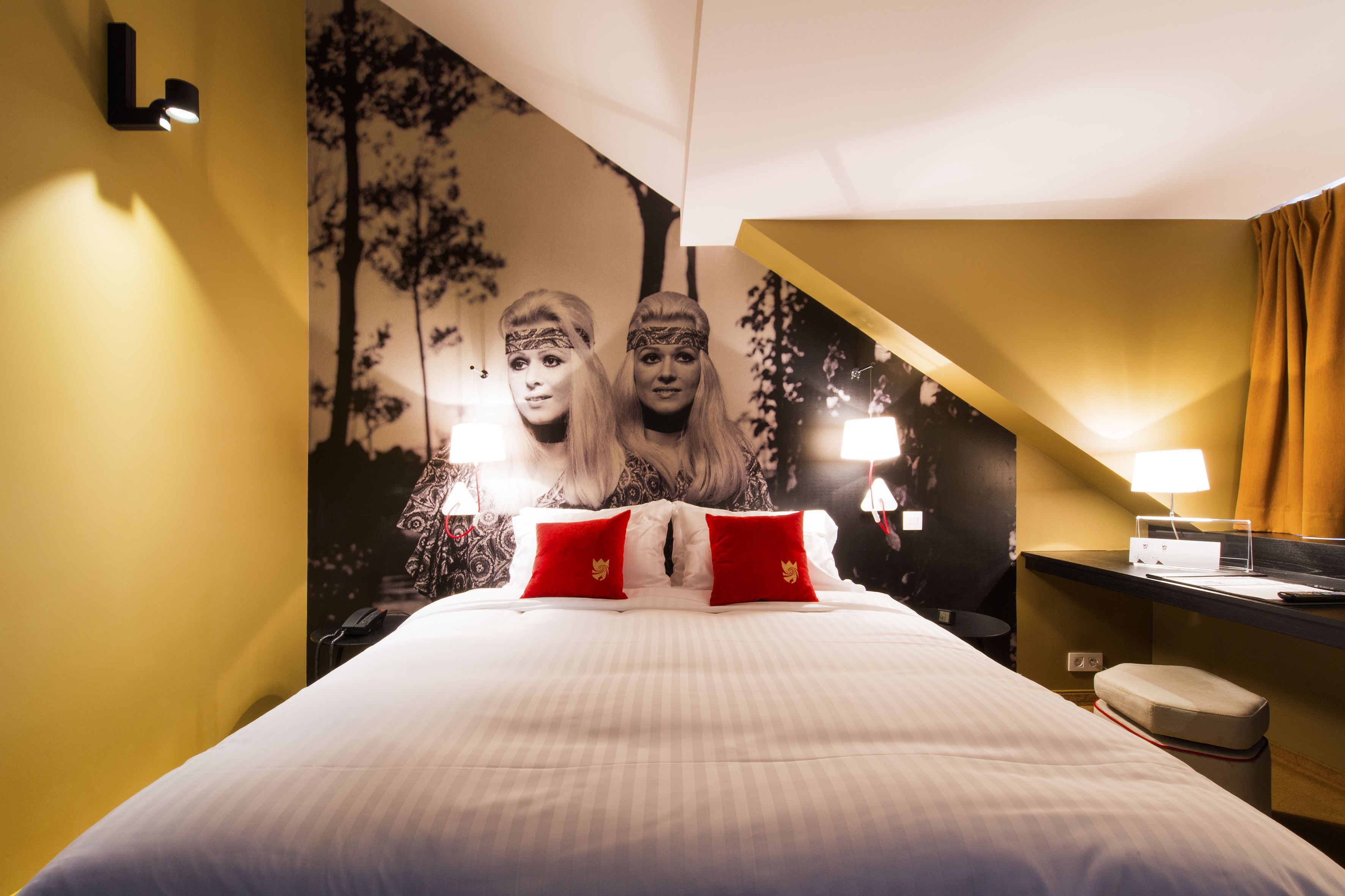 A hotel room with a bed, red pillows, a nightstand, a lamp, and a wall with a tree mural.
