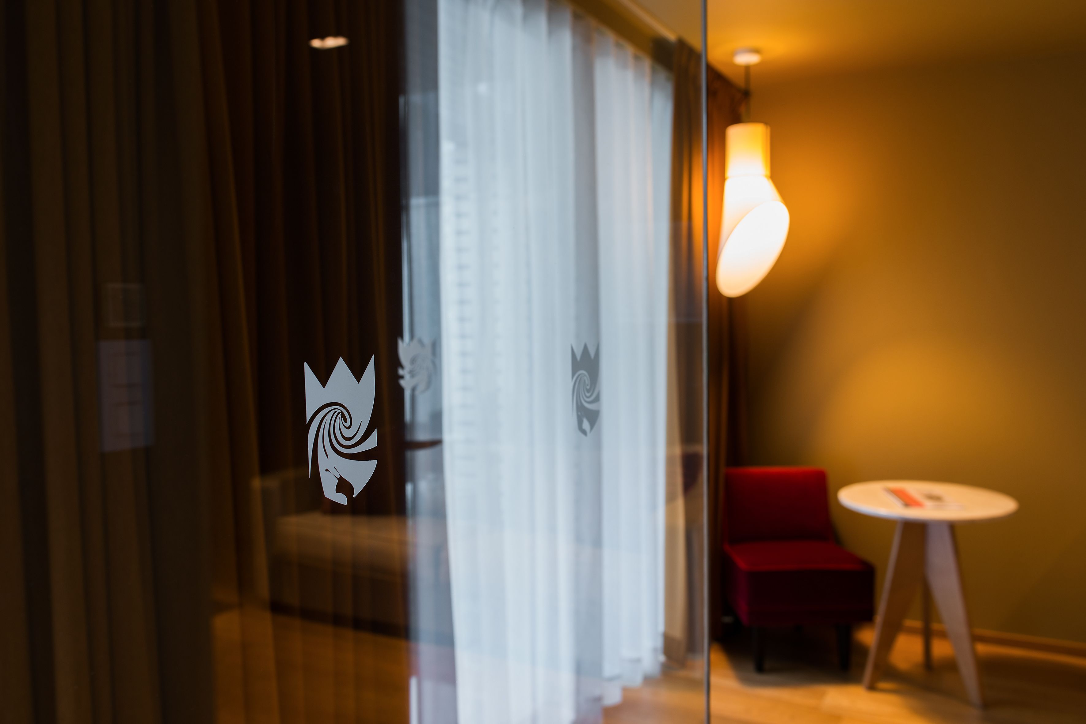 Habitación de hotel con mampara de vidrio, una silla roja, una mesita redonda y un aplique de pared.