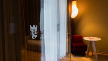A hotel room with a glass partition, a red chair, a small round table, and a wall-mounted light fixture.