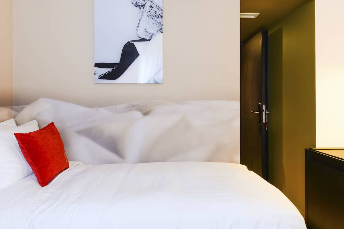 A neatly made bed with white linens and a red accent pillow, a black and white artwork on the wall, and a dark wooden wardrobe with a light switch.