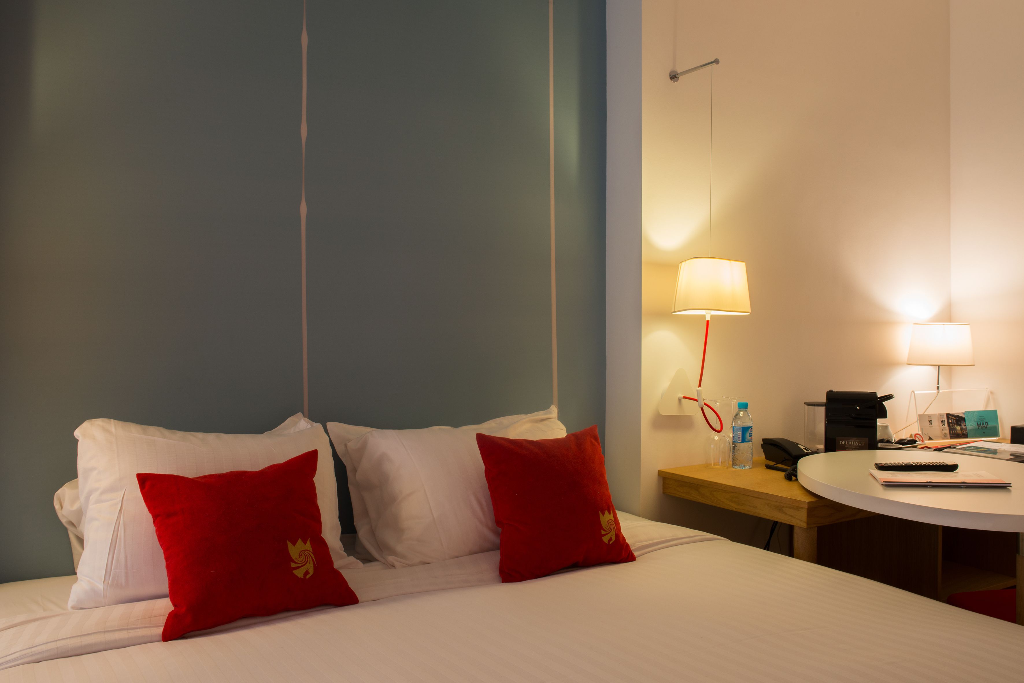 Habitación de hotel con cama, dos almohadas rojas, mesita de noche con lámpara, teléfono y una pequeña mesa redonda con control remoto y libreta.