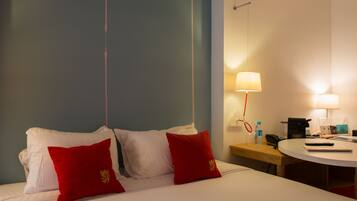Hotelkamer met een bed, twee rode kussens, een nachtkastje met een lamp, een telefoon en een klein rond tafeltje met een afstandsbediening en een notitieboekje.