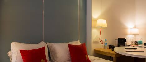 Habitación de hotel con cama, dos almohadas rojas, mesita de noche con lámpara, teléfono y una pequeña mesa redonda con control remoto y libreta.