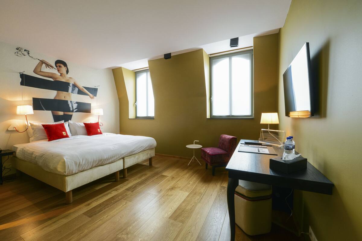 A hotel room with a bed, bedside lamps, a desk with a chair, a small table, and a TV mounted on the wall.