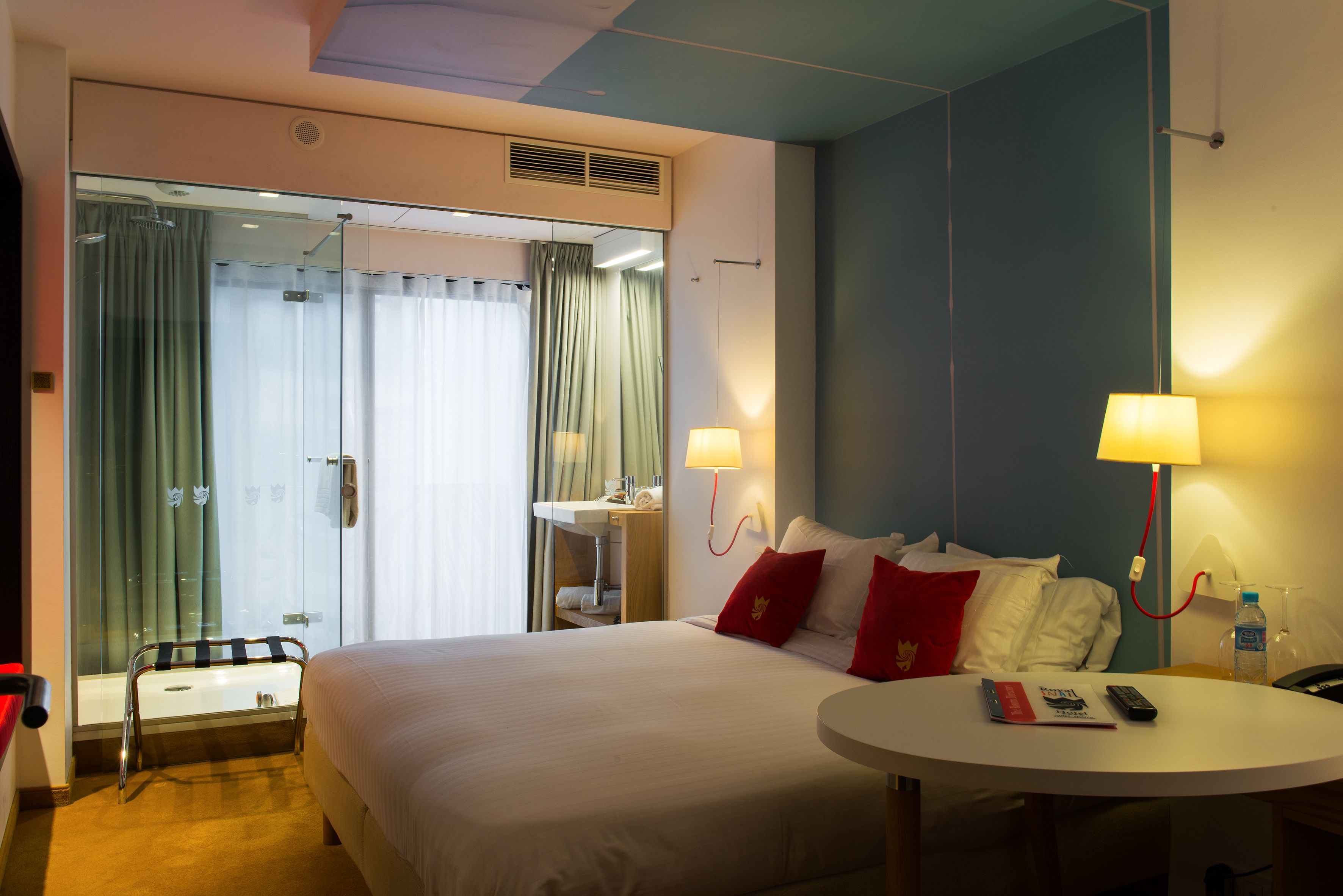 A hotel room with a large bed, two bedside lamps, a round table, a small bedside table, a mirror, and a window with curtains.