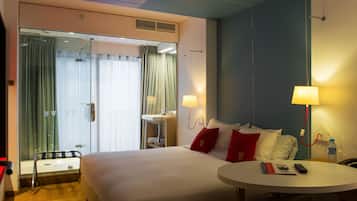Habitación de hotel con una cama grande, dos lámparas de noche, una mesita redonda, una mesita auxiliar pequeña, un espejo y una ventana con cortinas.