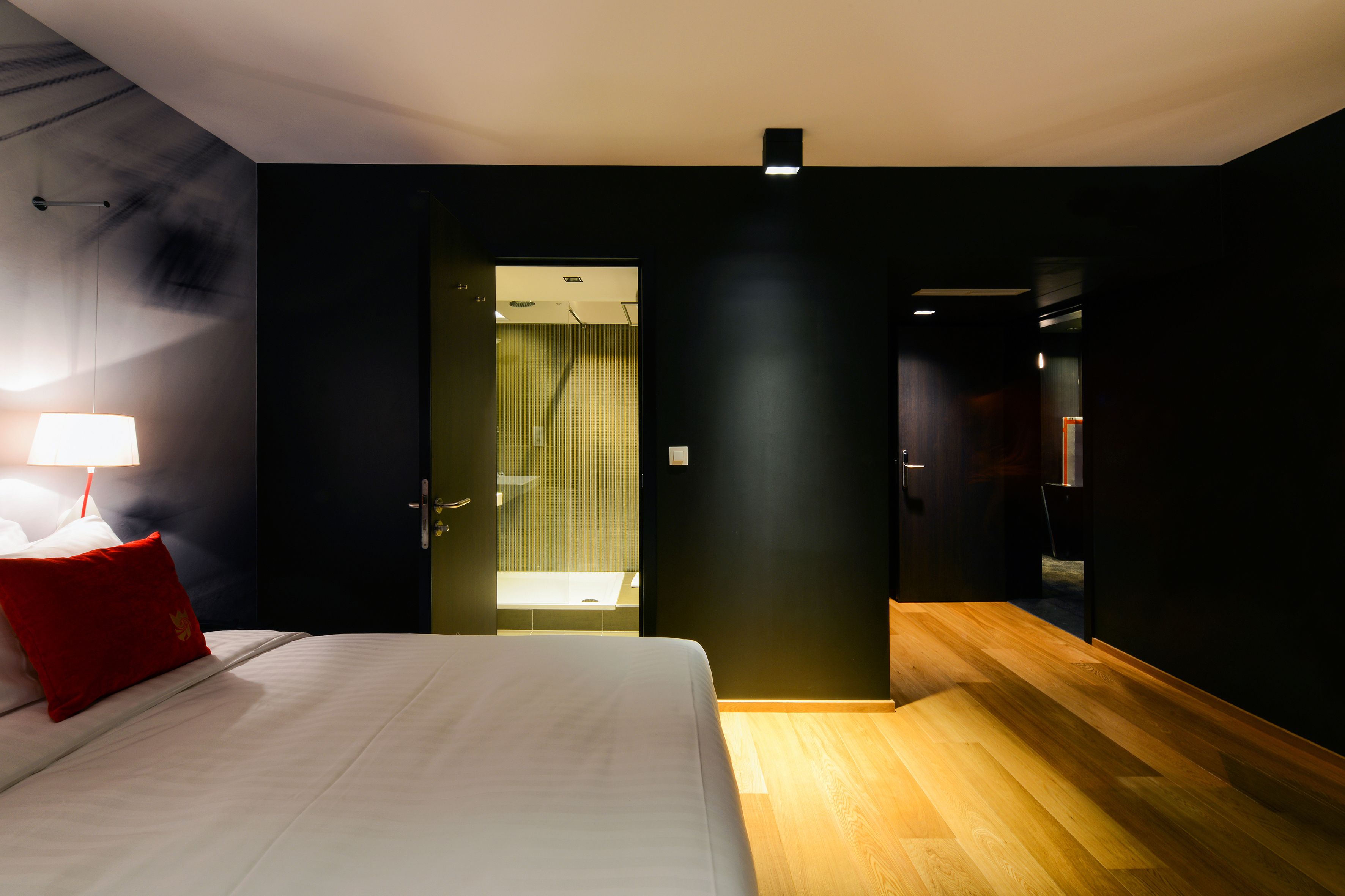 A modern hotel room with a bed, a red pillow, a nightstand with a lamp, and a wooden floor.