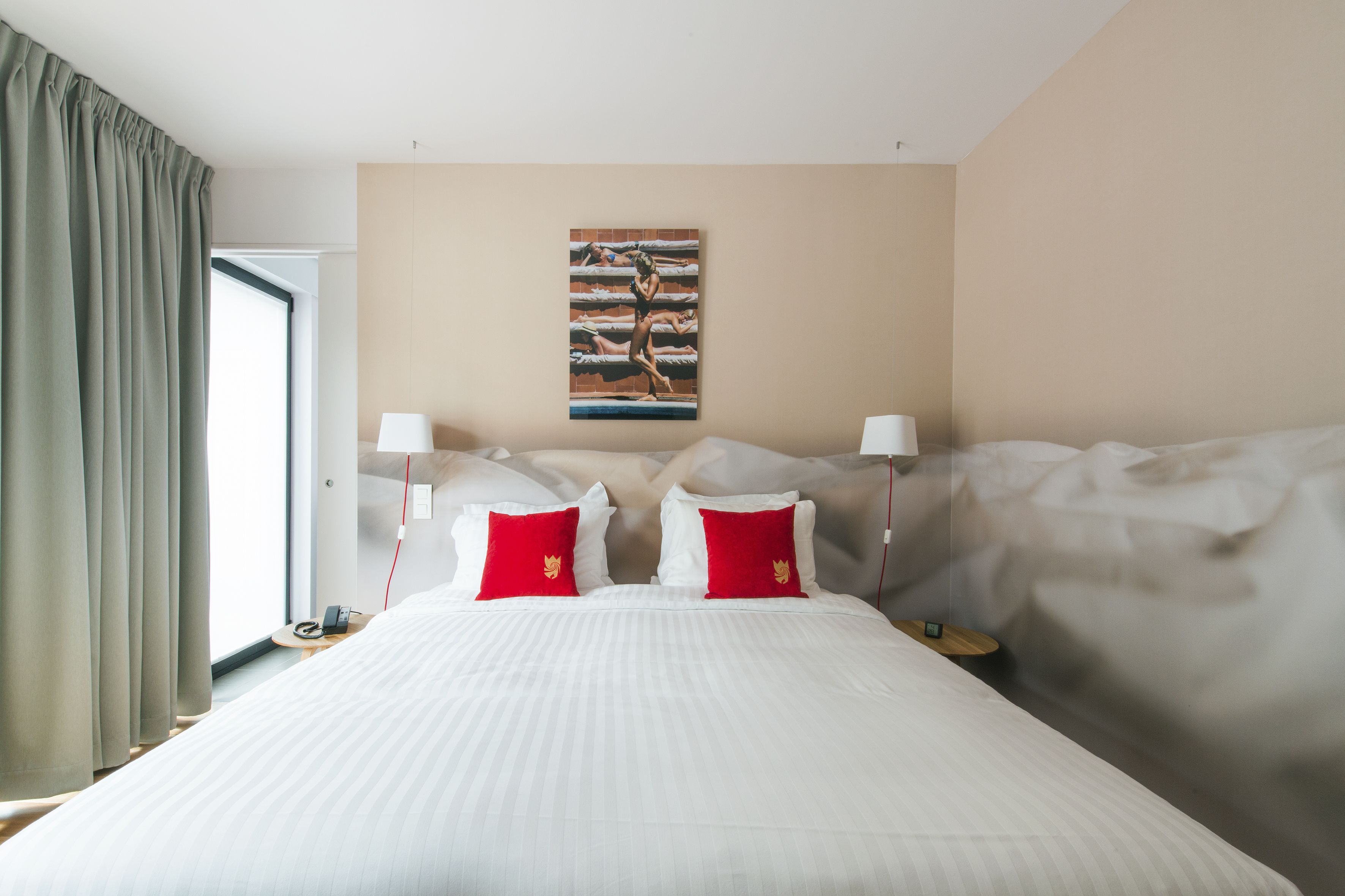 A hotel room with a large bed, two red pillows, a painting on the wall, and a window with curtains.