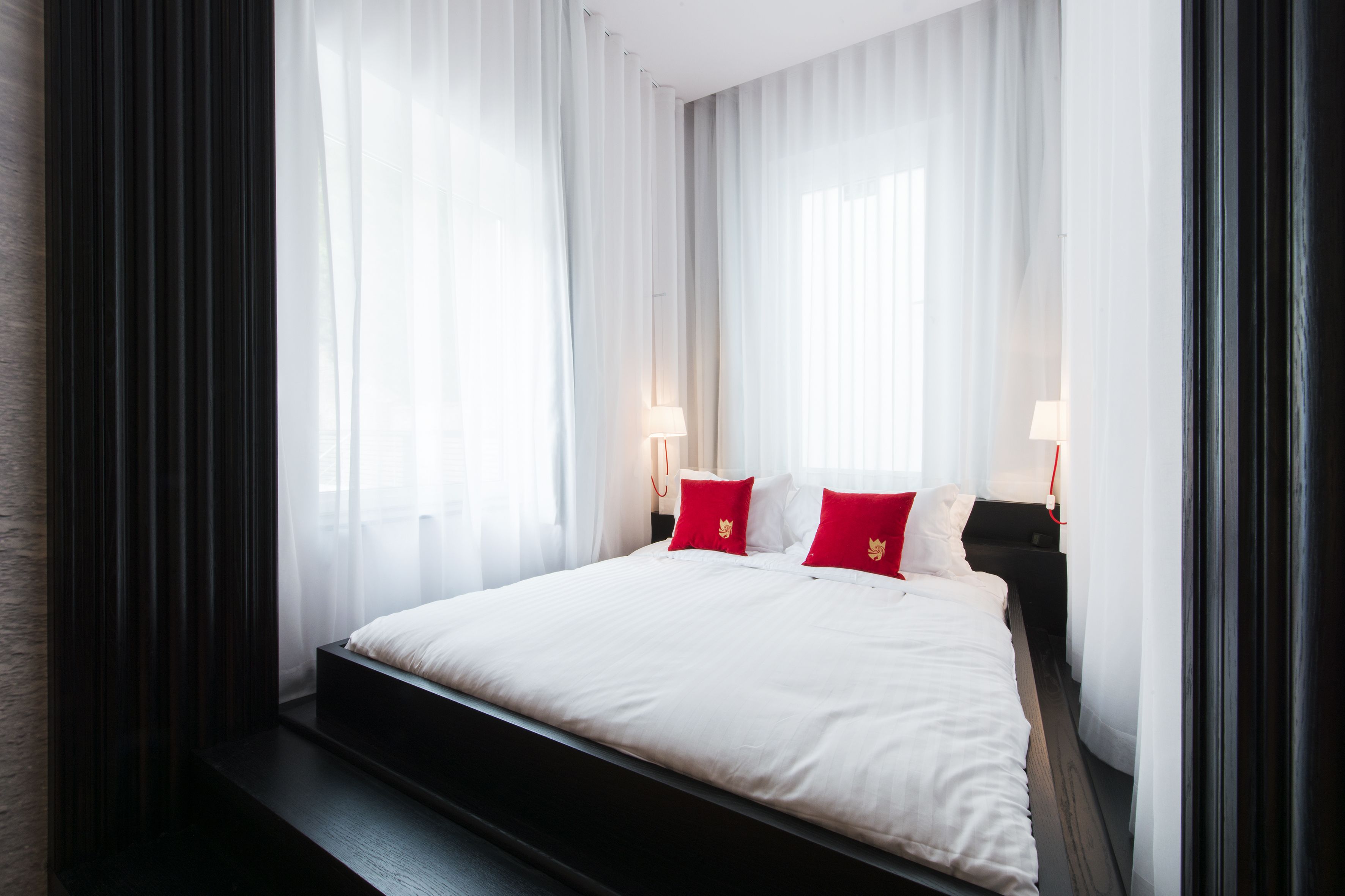 Una habitación de hotel con una cama grande, ropa de cama blanca, almohadas rojas y cortinas translúcidas.