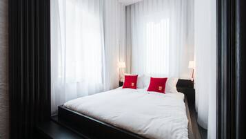 Una habitación de hotel con una cama grande, ropa de cama blanca, almohadas rojas y cortinas translúcidas.
