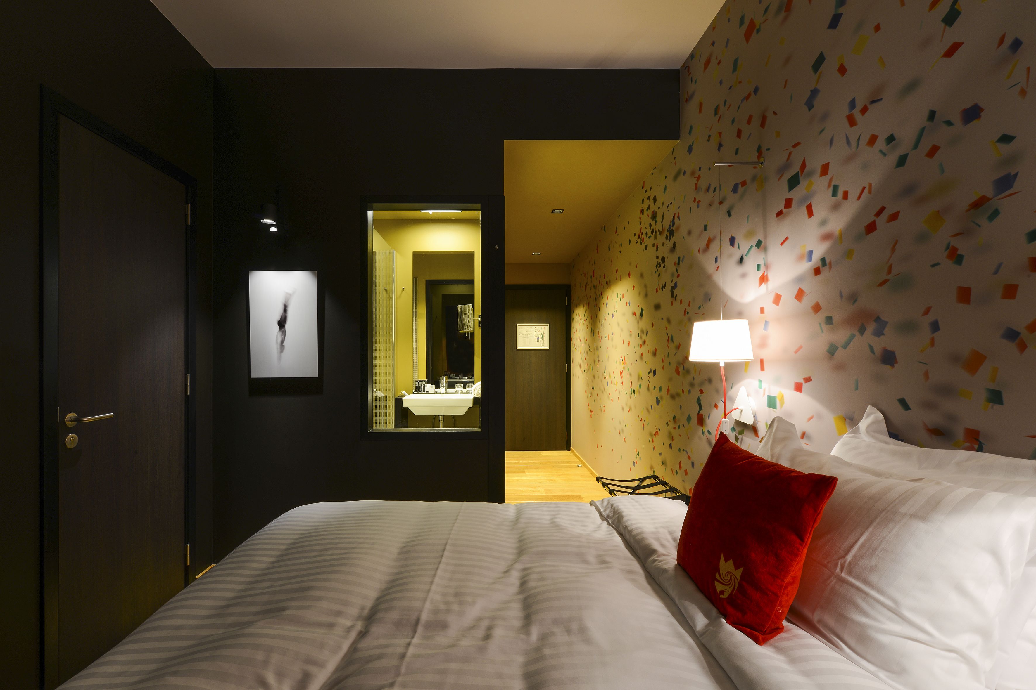 A hotel room with a bed, a red pillow, a mirror, and a wall with colorful square patterns.