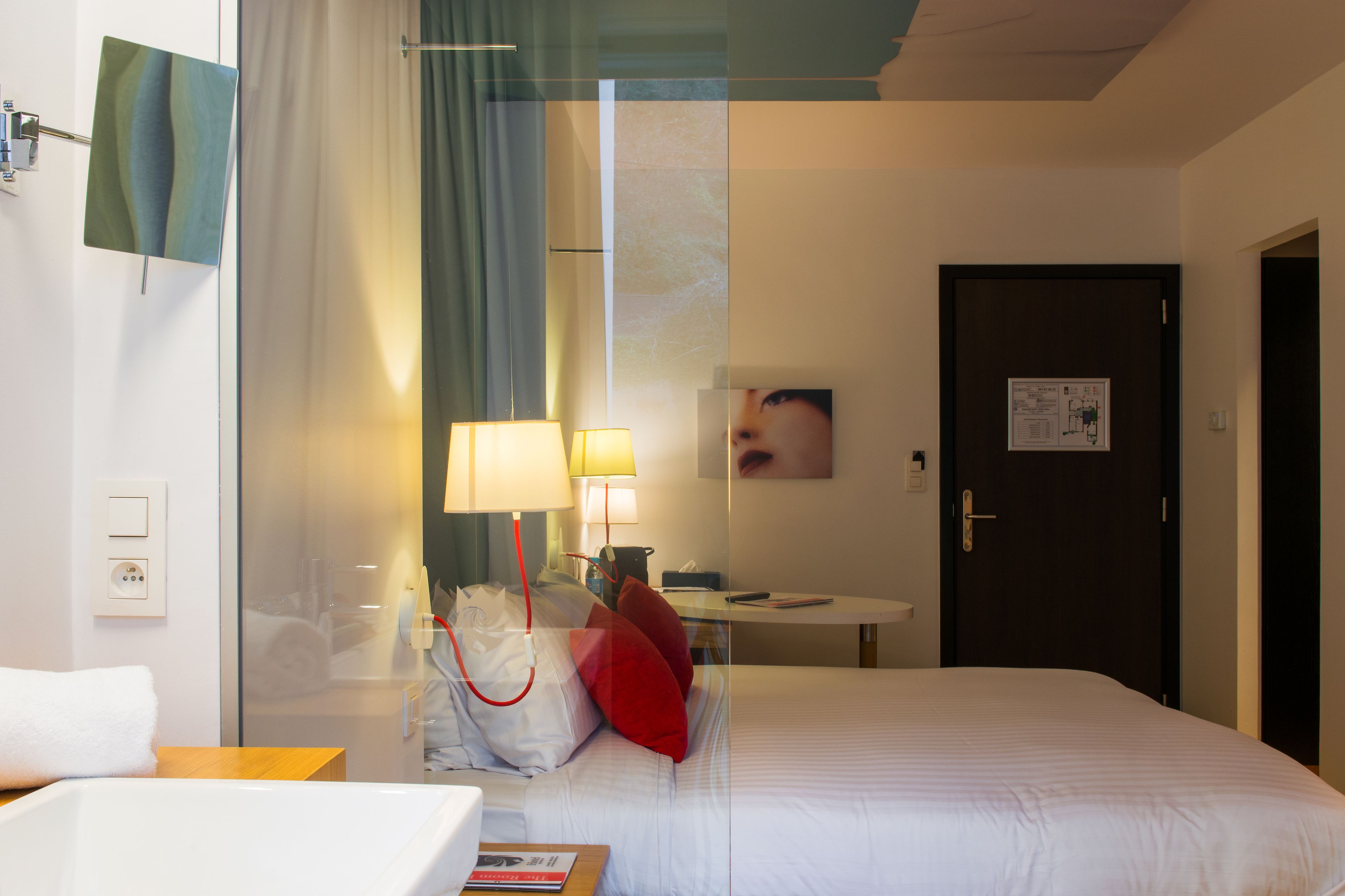A hotel room with a bed, bedside lamps, a glass shower enclosure, a bathroom with a sink, and a door with a poster.