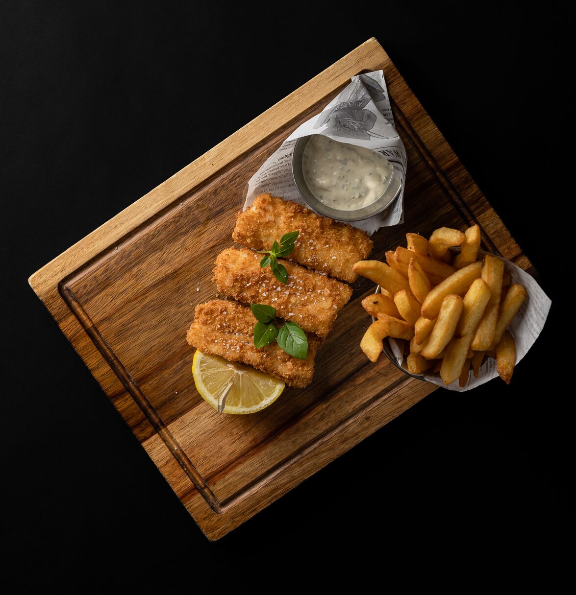 Una tabla de madera con filetes de pescado rebozado, papas fritas y una rodaja de limón.