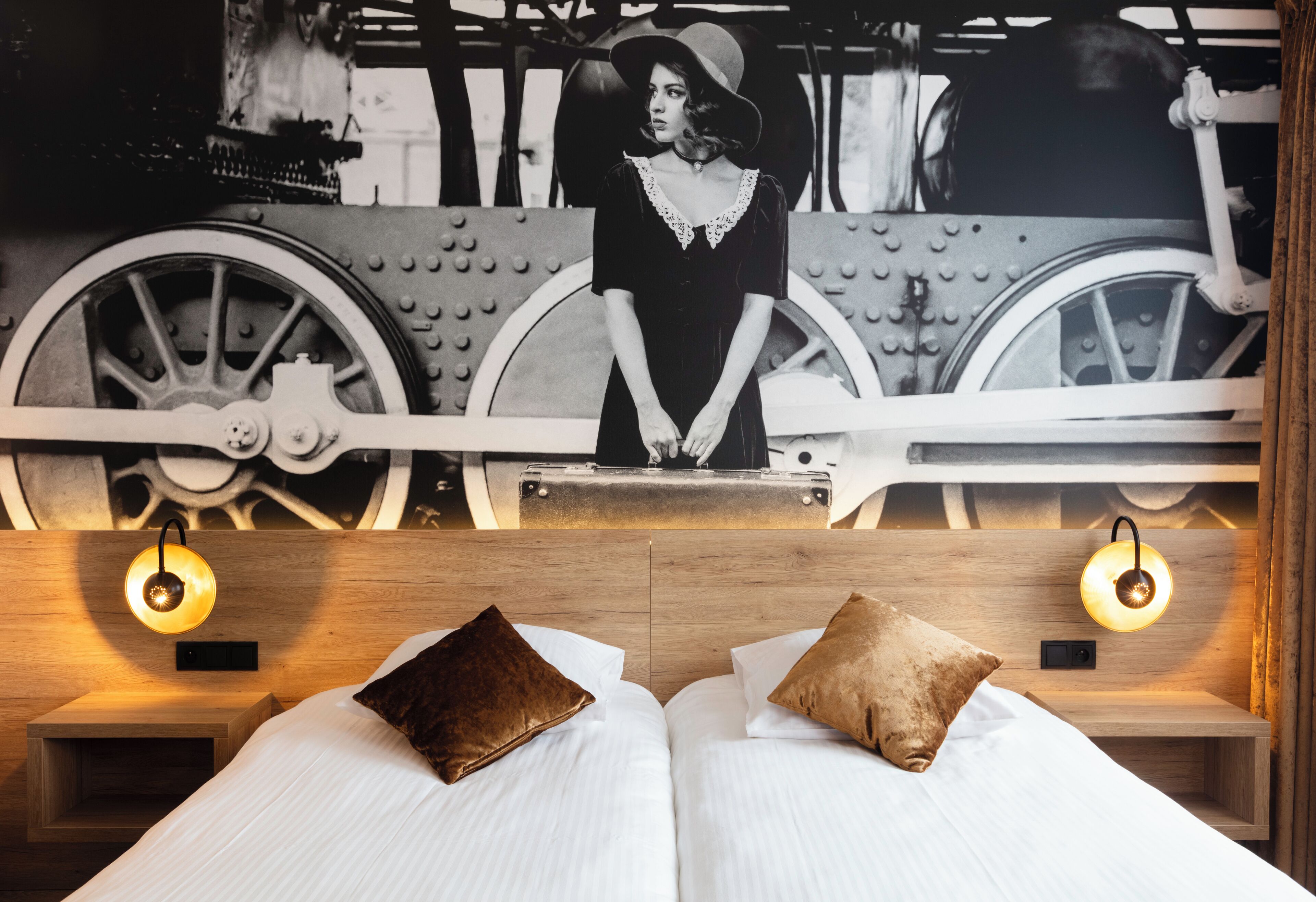 A bed with two pillows, a headboard with a wall art of a woman and a vintage train, and two wall-mounted light fixtures.