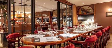 A long dining table with red upholstered chairs, a view of the kitchen through large windows, and a chandelier.