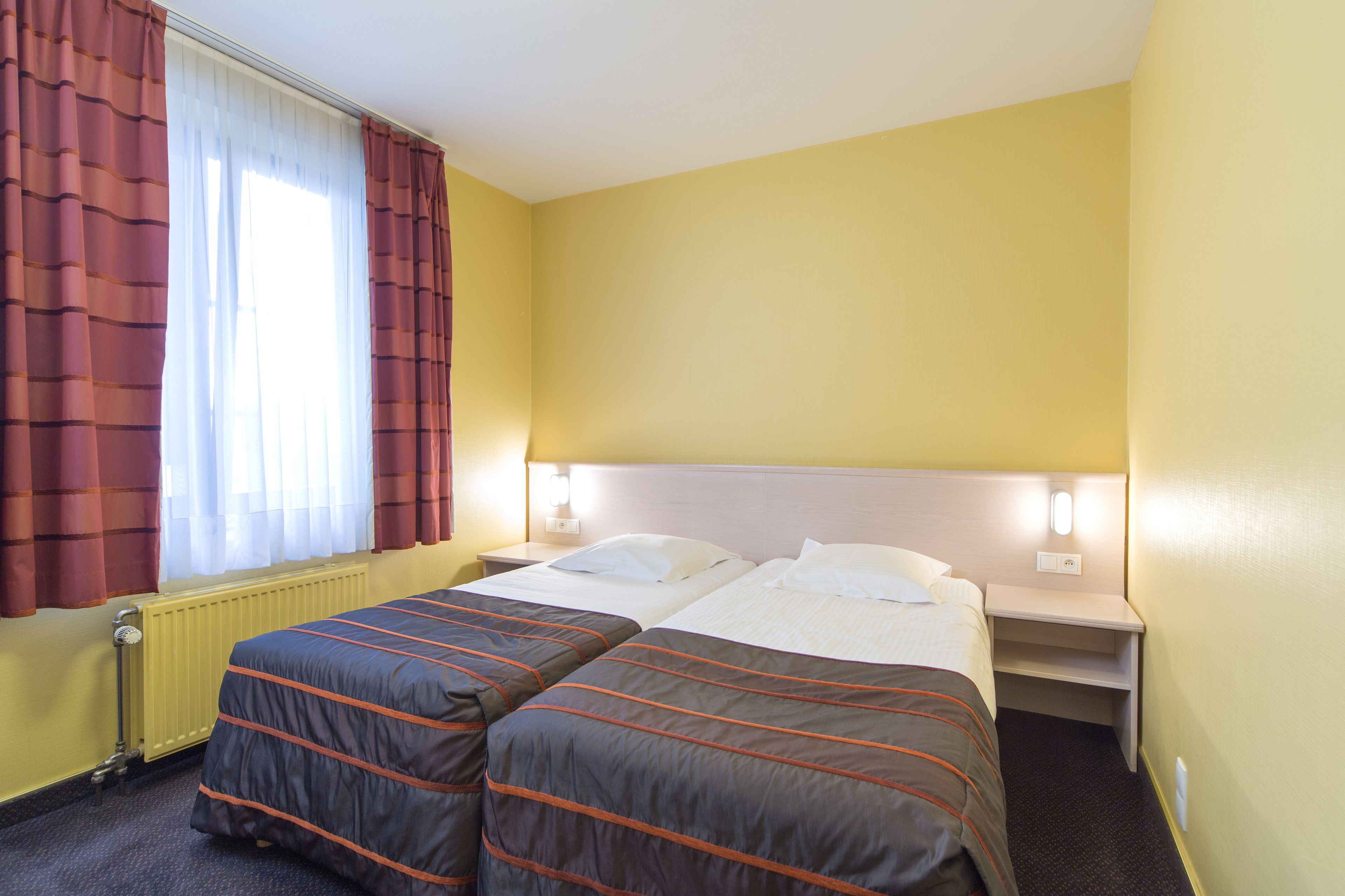 A hotel room with two beds, a window with curtains, a radiator, and bedside lamps.