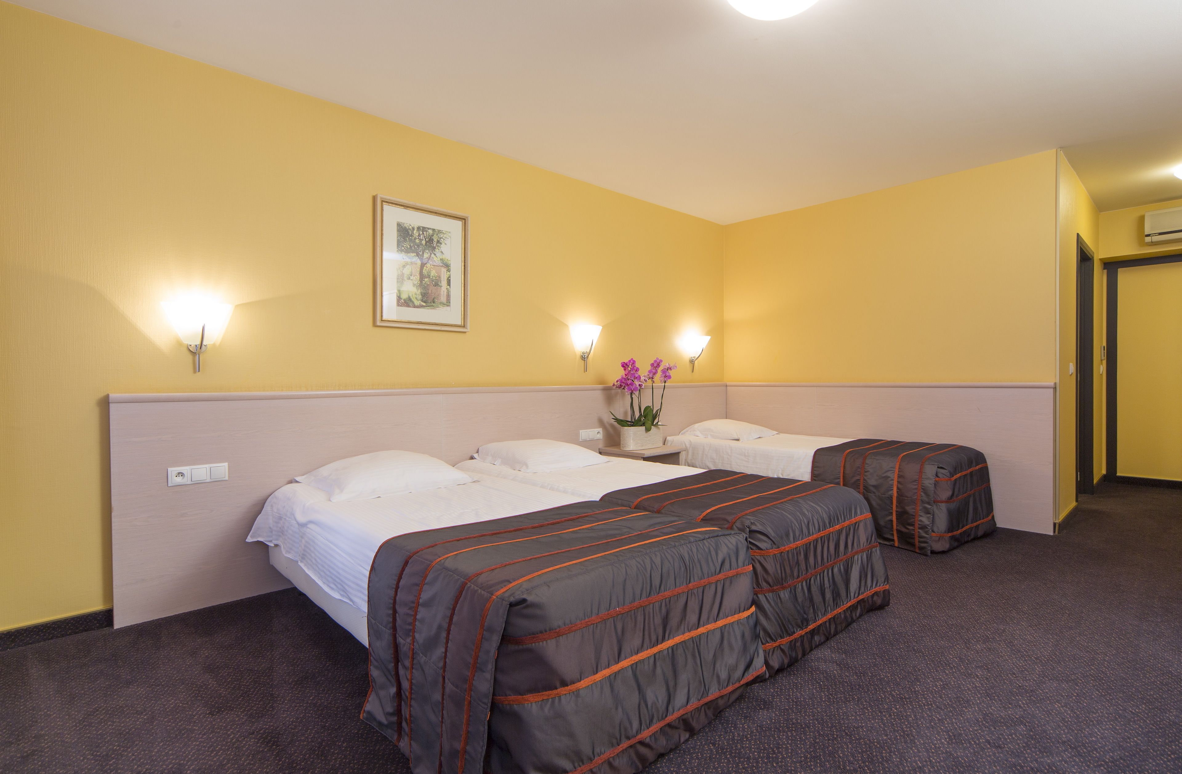 A hotel room with two beds, a nightstand with a vase of flowers, and wall-mounted lights.