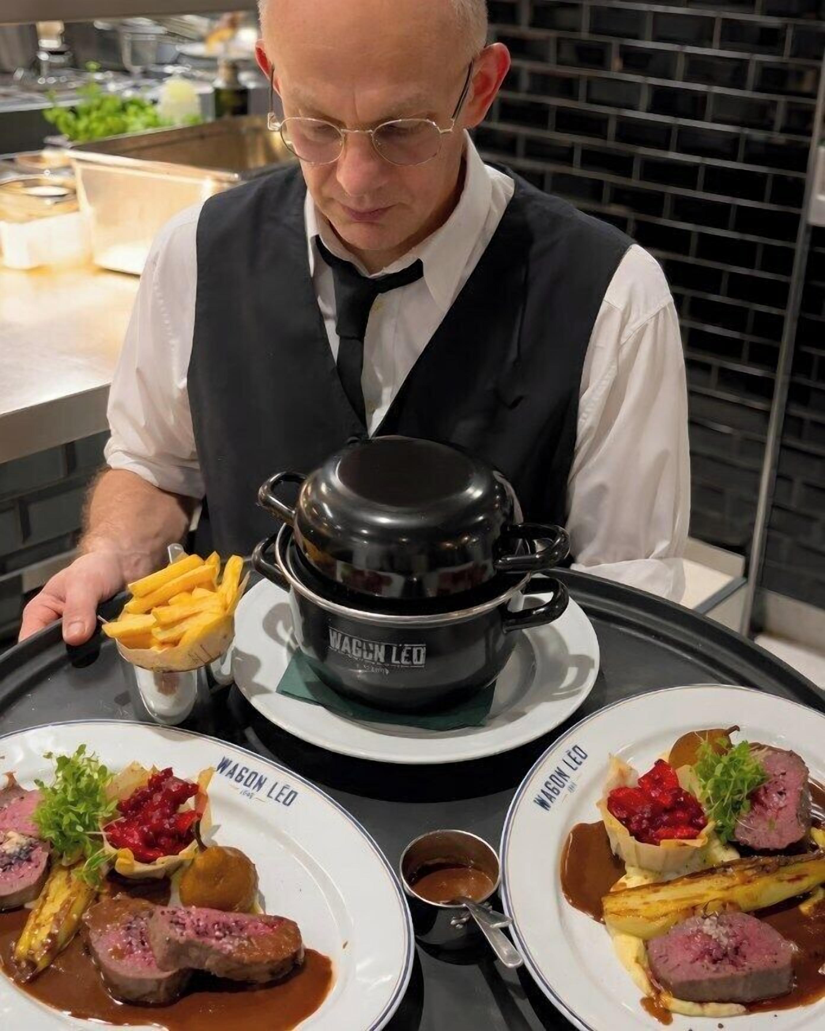 Ein Koch bereitet Essen in einer Küche zu, wobei ein Topf mit der Aufschrift 'MASON LED' und Teller mit Speisen, darunter Steak und Pommes Frites, zu sehen sind.