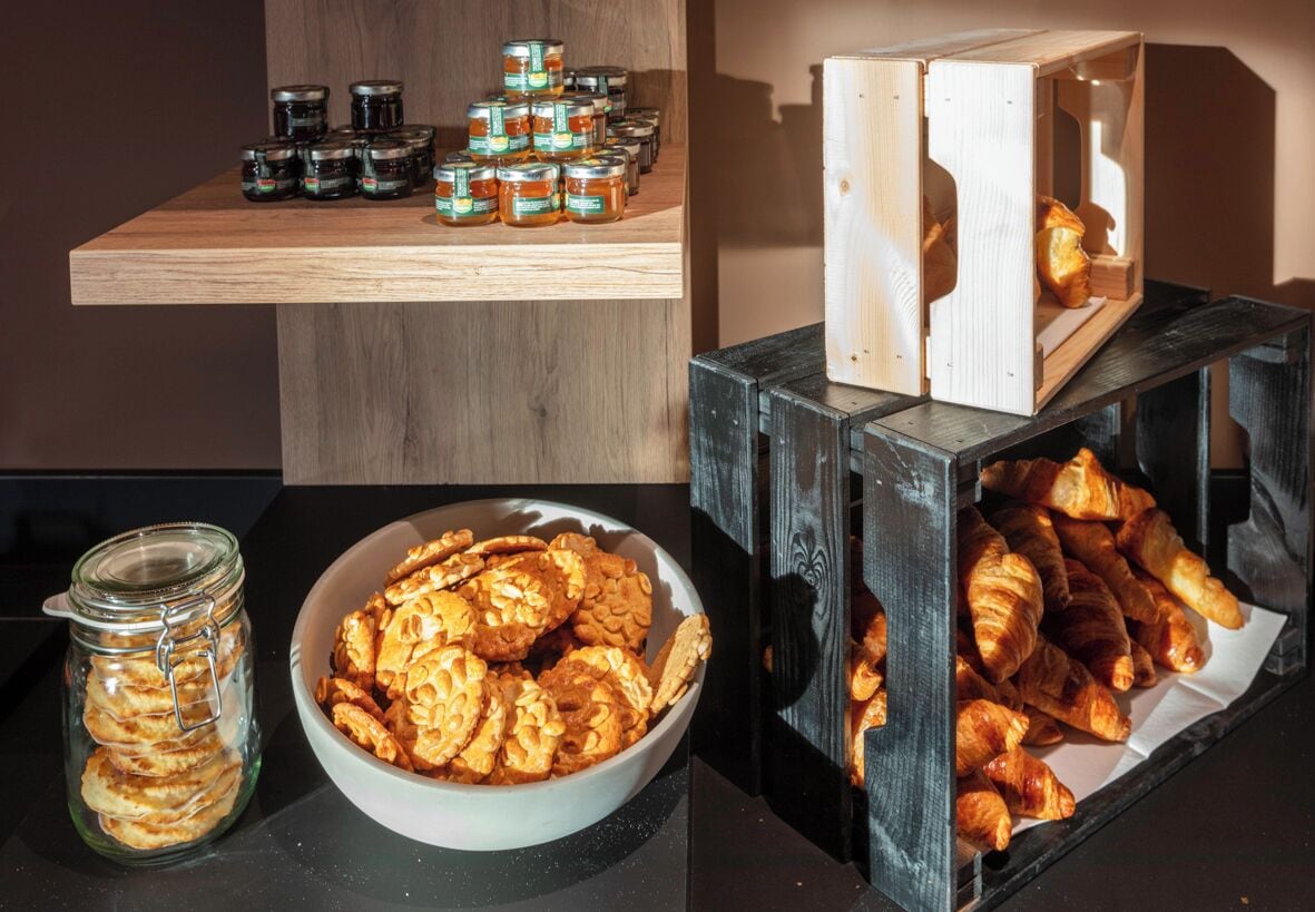 En træhylde med syltetøjsglas, en skål med småkager og et udstilling af croissanter i en trækasse.