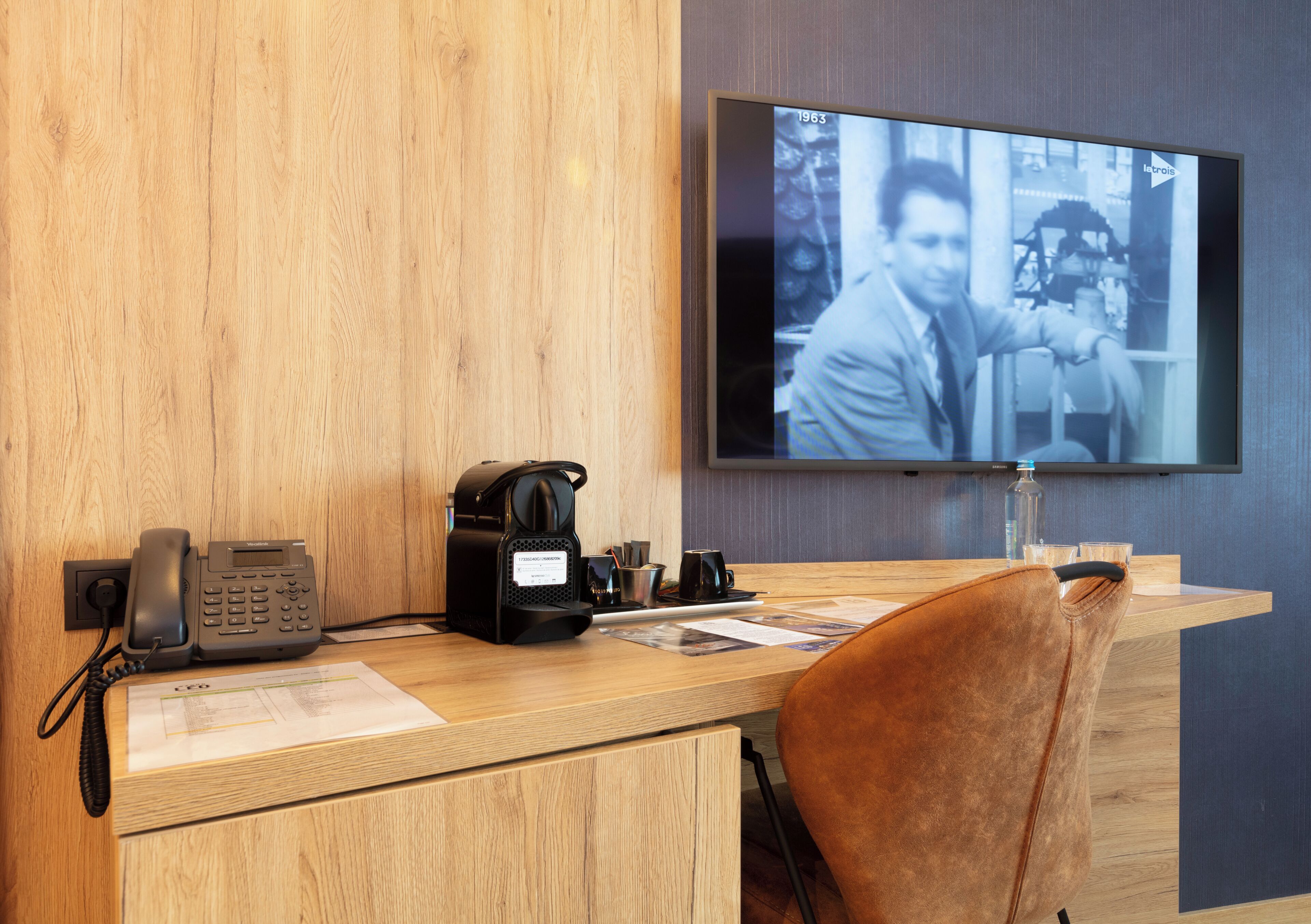A wooden desk with a telephone, coffee maker, and papers, a brown leather chair, and a television displaying a blurred image.