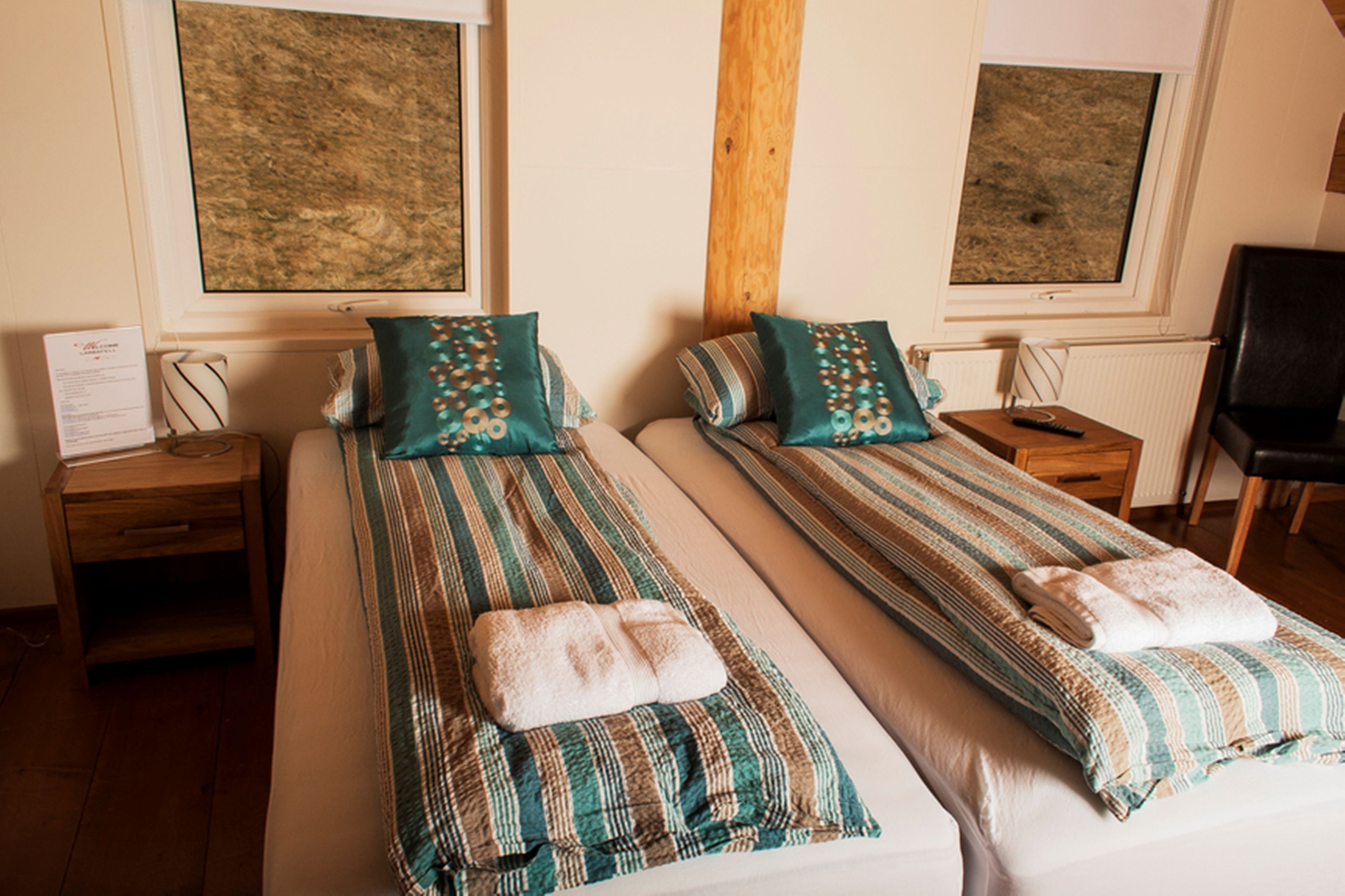 Habitación doble con dos camas, cada una con ropa de cama a rayas y almohadas a juego. Cuenta con cabeceras de madera, una mesita de noche con lámpara y una ventana con persianas.