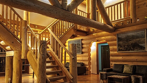 Interior de una cabaña de troncos con una escalera de madera, una sala de estar con sofá y reposapiés, y un cuadro grande enmarcado en la pared.