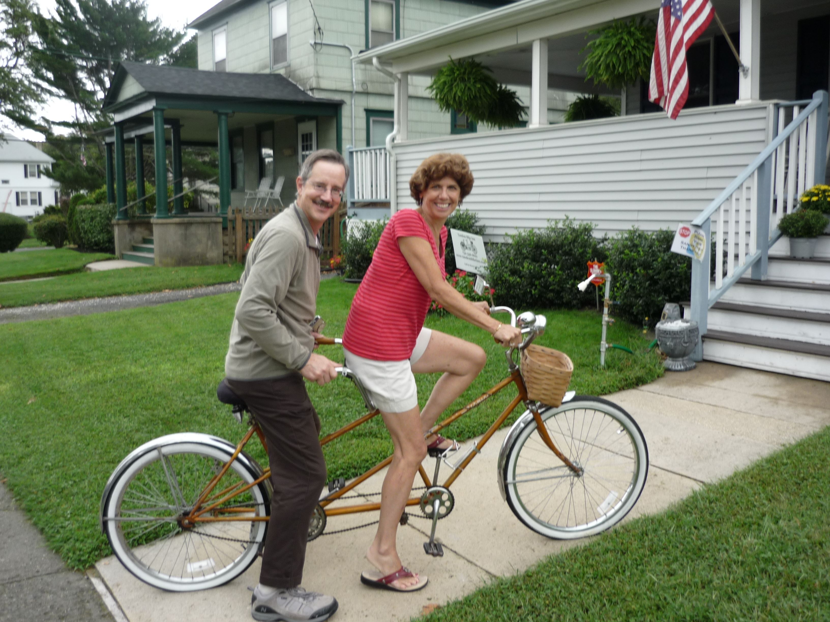 Ein Mann und eine Frau fahren mit dem Fahrrad vor einem Haus mit Veranda und einer amerikanischen Flagge.