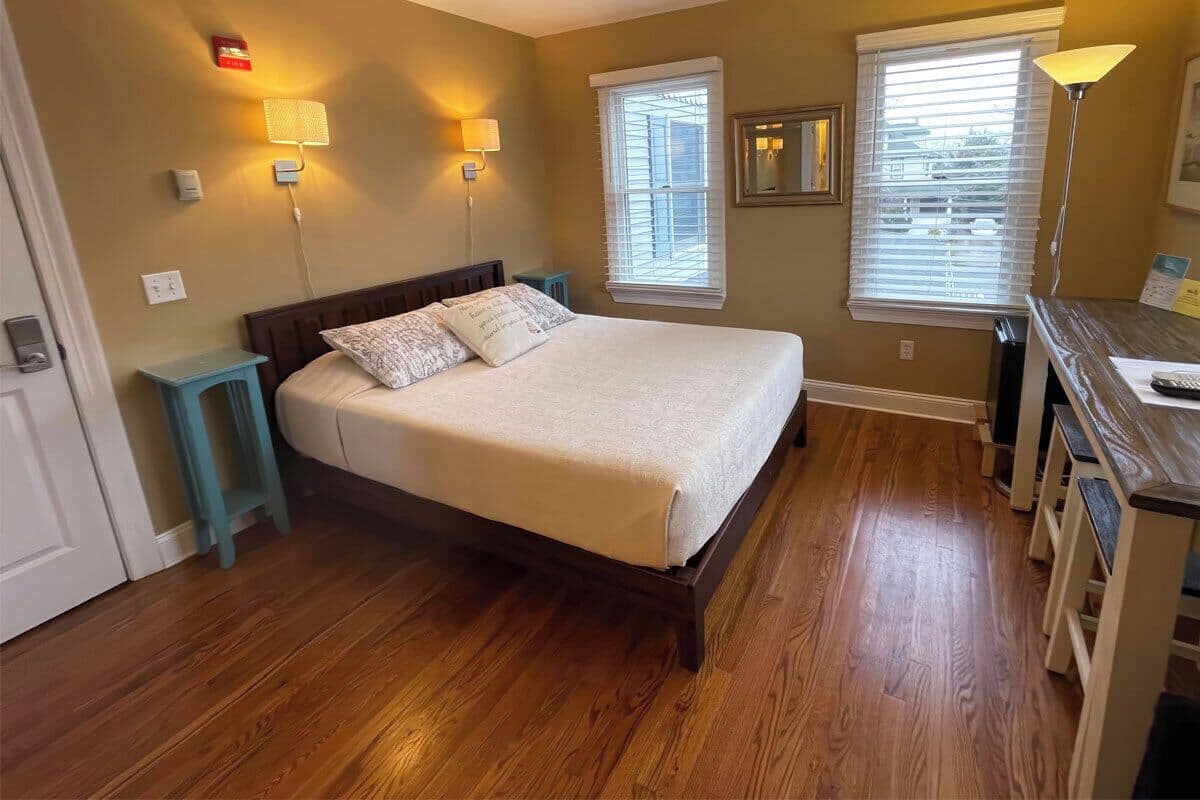A bedroom with a bed, two bedside lamps, a nightstand, a desk with a chair, and a window with blinds.