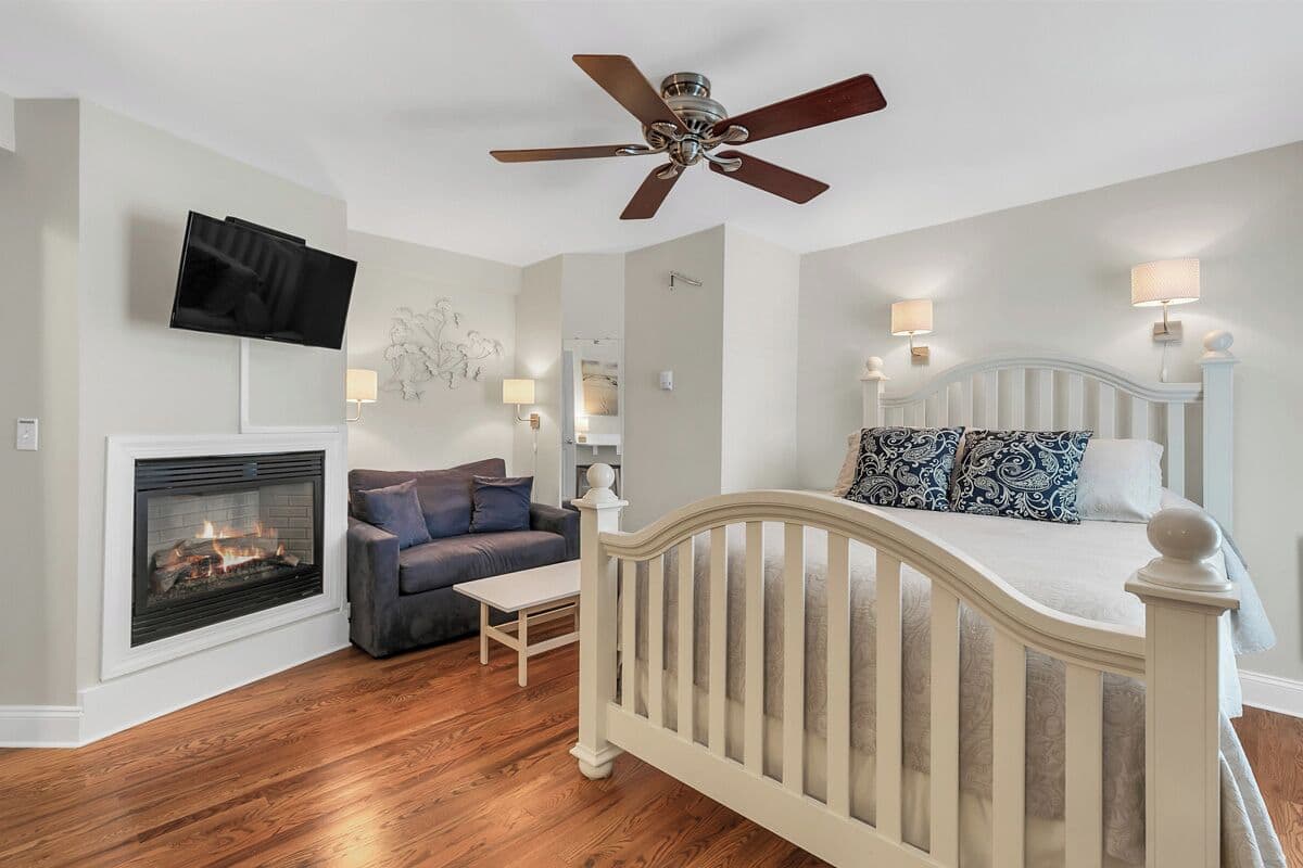 A bedroom with a white bed, a ceiling fan, a fireplace, a television, and a sofa.