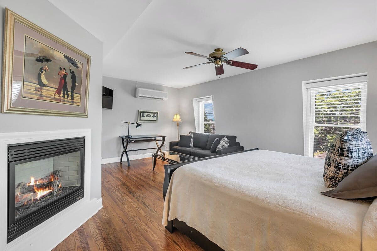 A bedroom with a fireplace, a bed, a ceiling fan, a window with blinds, and a painting on the wall.