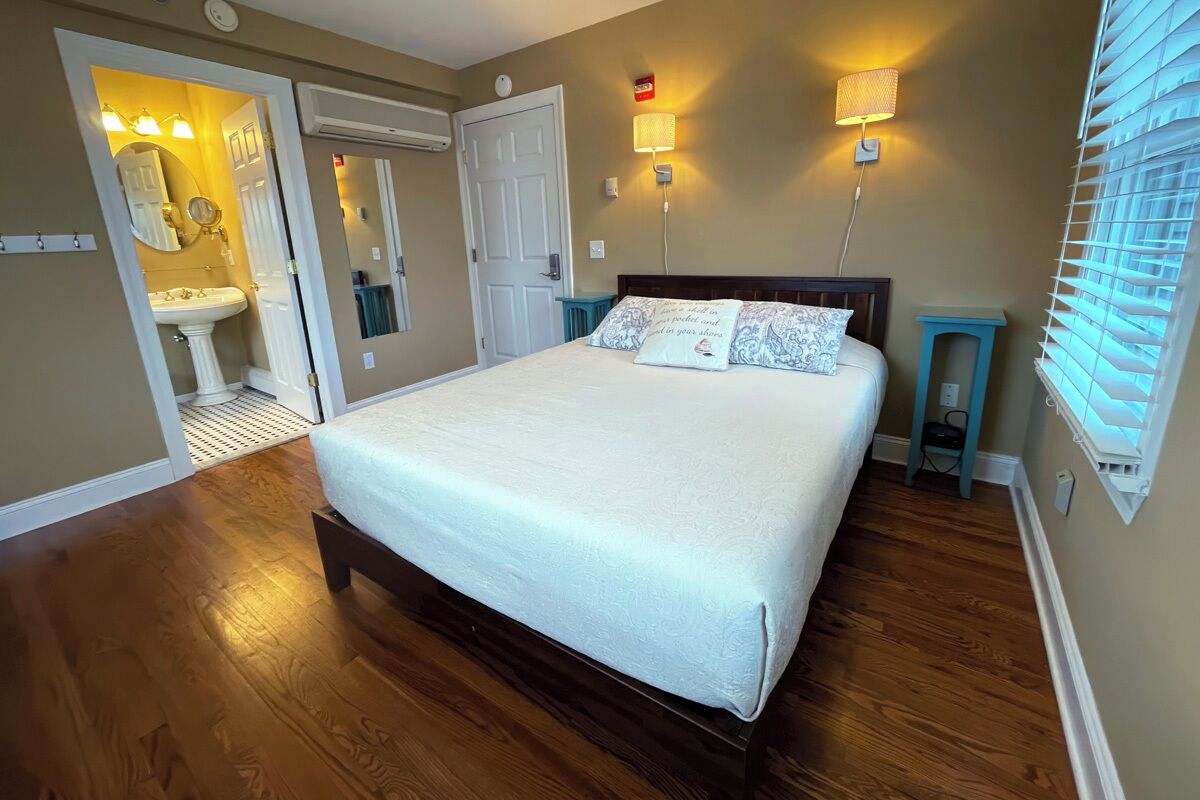 Un dormitorio con cama, mesitas de noche, un tocador, una ventana con persianas, una puerta y un baño con lavamanos y espejo.