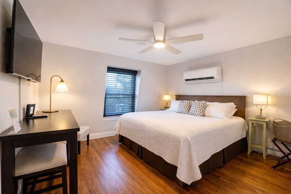 A hotel room with a bed, desk, chair, ceiling fan, window with blinds, bedside lamps, and an air conditioning unit.