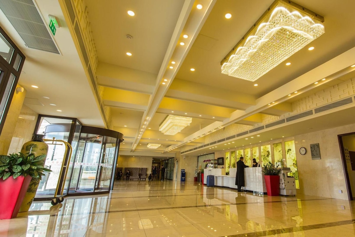 A hotel lobby with a reception desk, potted plants, a glass elevator, and a polished floor.