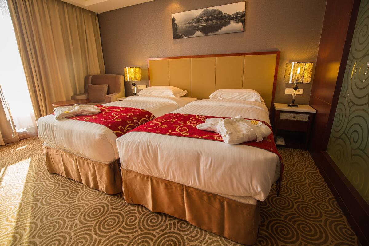 A hotel room with two beds, a chair, lamps, and a painting on the wall.