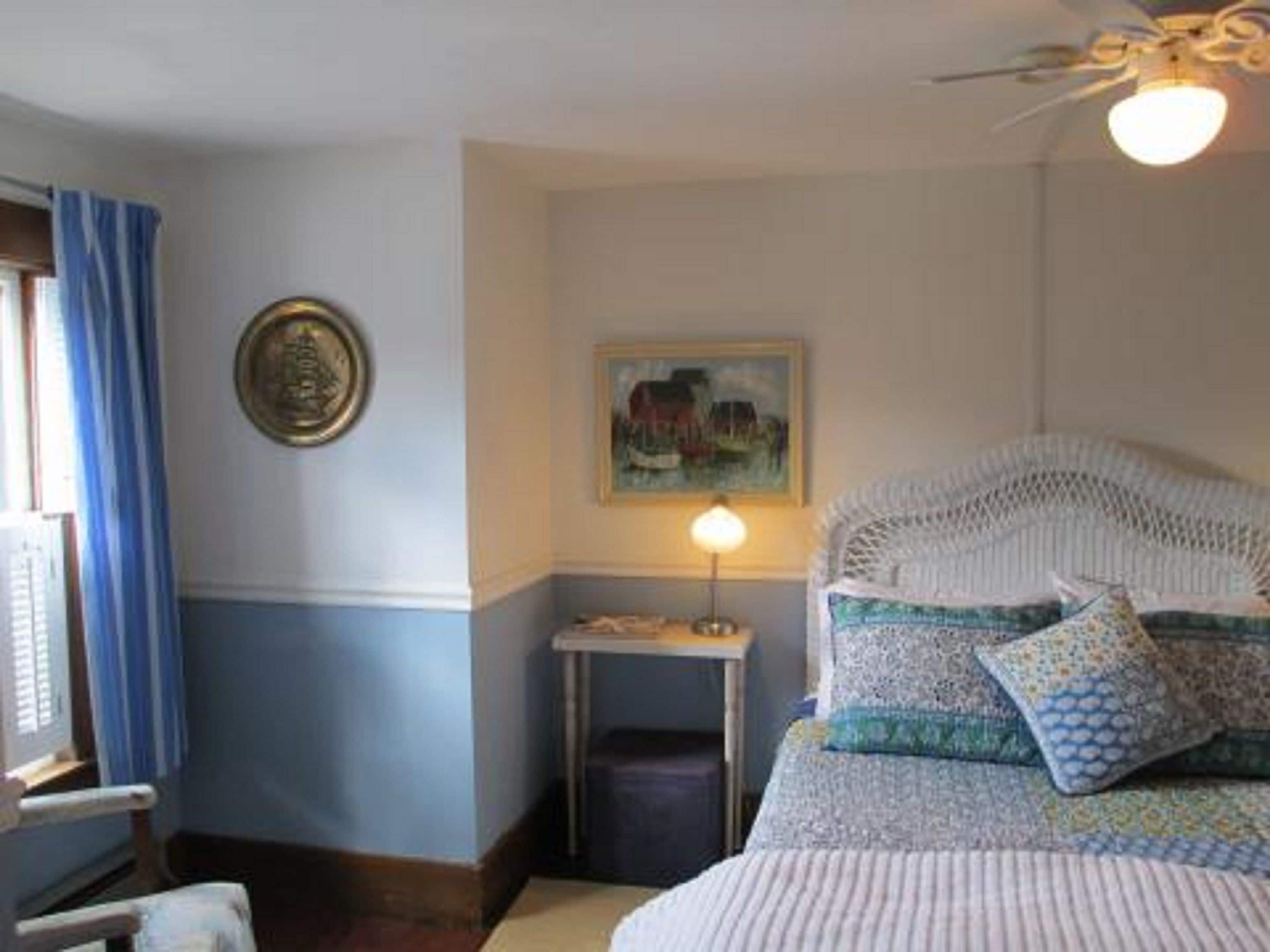 A bedroom with a bed, a nightstand, a lamp, a painting, and a ceiling fan.