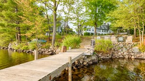 A wooden dock leading to a stone staircase with a view of a white house and green chairs by a lake.