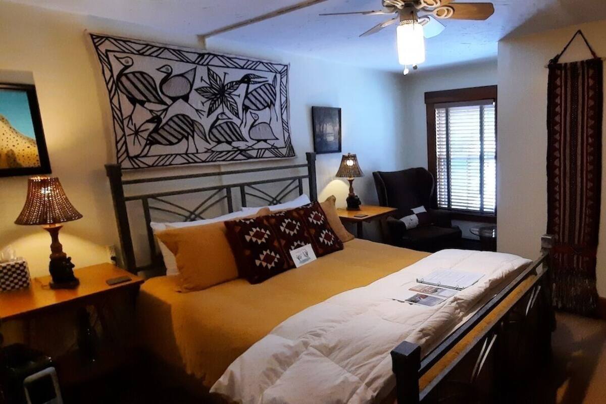 A hotel room with a bed, a desk with a lamp, a chair, a window with blinds, and a decorative wall piece.
