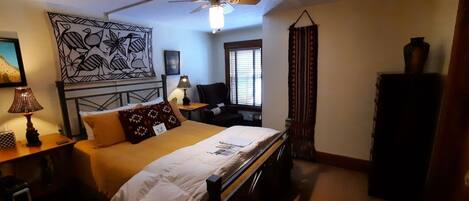 A hotel room with a bed, a desk with a lamp, a chair, a window with blinds, and a decorative wall piece.
