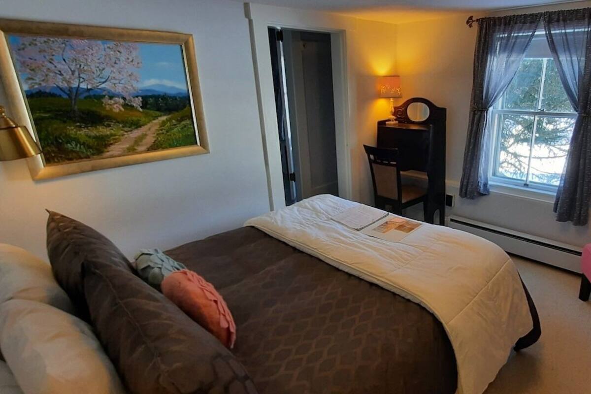 A bedroom with a bed, a chair, and a desk. There is a painting on the wall and a window with curtains.