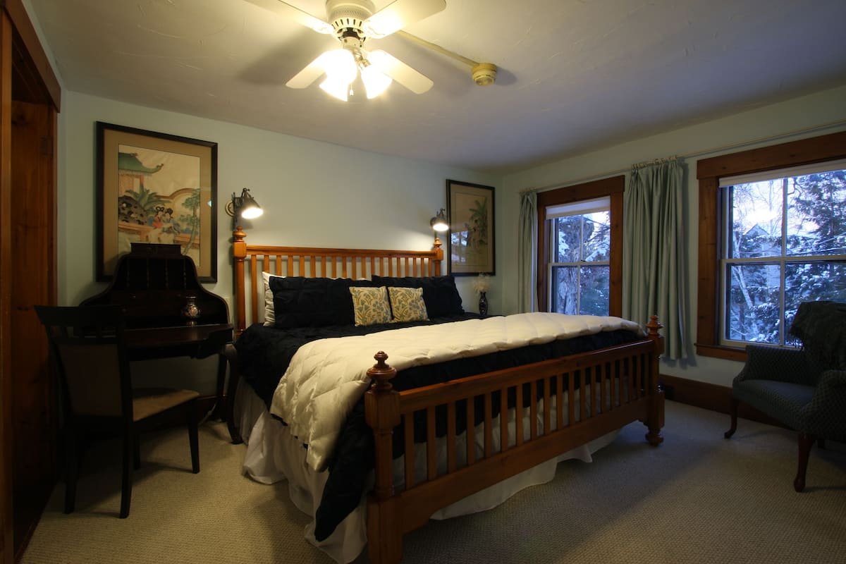 A bedroom with a wooden bed, a desk, and a chair. There are two windows with curtains, and a ceiling fan is visible.