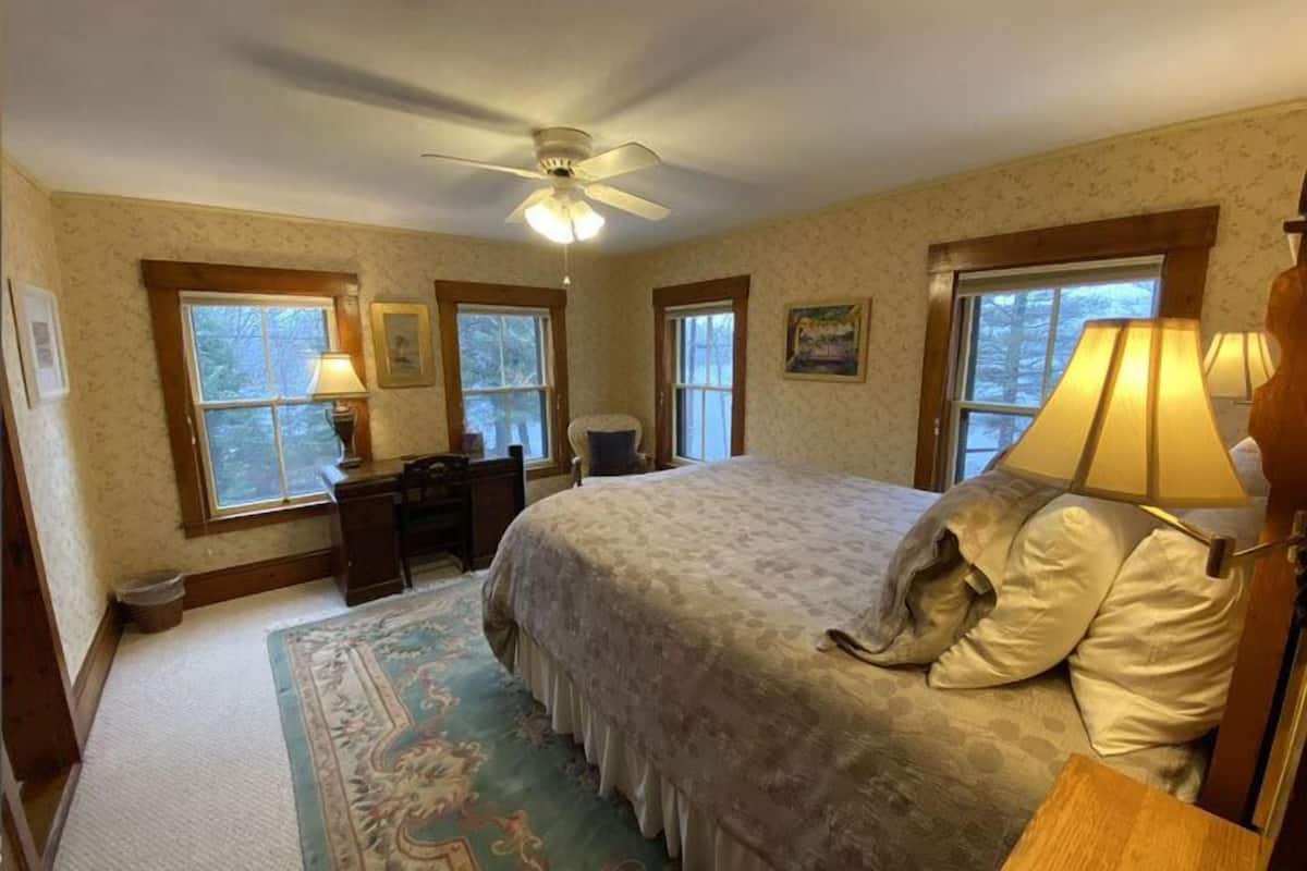 A bedroom with a large bed, two windows, a desk, and a ceiling fan.