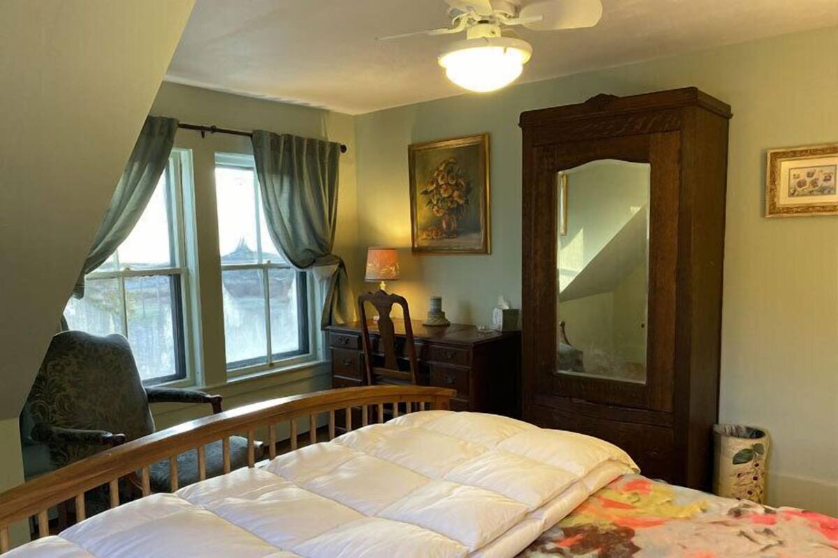 A bedroom with a bed, a chair, a desk, a mirror, and a window with curtains.