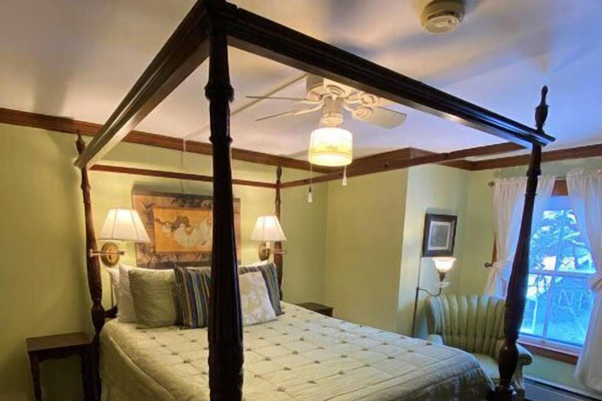 A four-poster bed with a canopy, a nightstand, a lamp, and a window with curtains.
