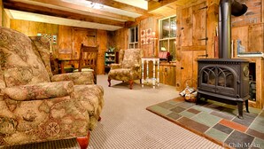 A cozy room with wooden walls, a large armchair, a smaller chair, a table, and a wood-burning stove.