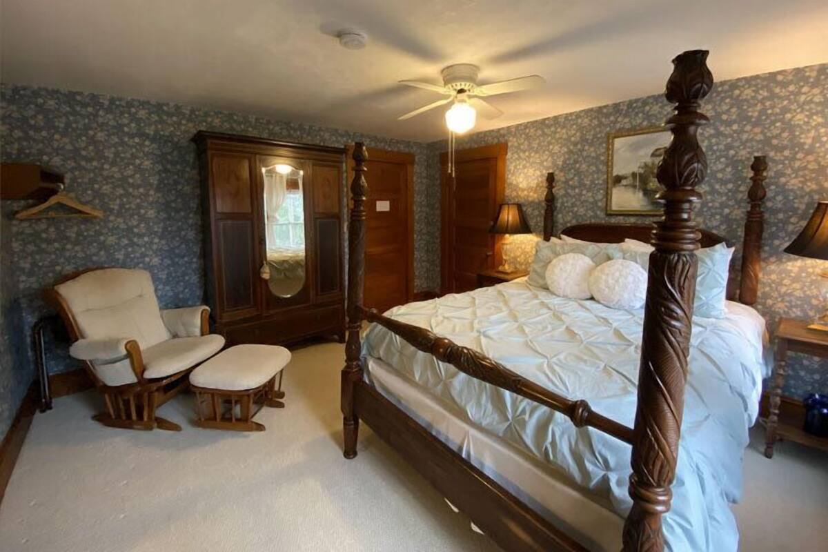A bedroom with a four-poster bed, a wooden wardrobe, a chair, and a ceiling fan.